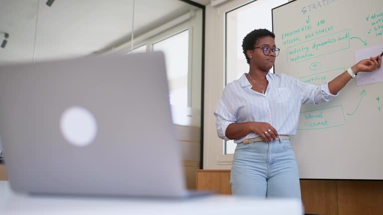 Confident young african woman conducts webinar, business training online. Online coach stands near flip chart in front of laptop and explains something to online audience